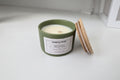 Candle in a green container with a wooden lid on a white surface, featuring 'AURA & ATLAS' branding.